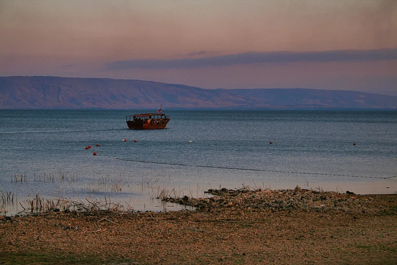A wooden boat resting on the calm waters of the Sea of Galilee at dusk with the Golan Heights visible across the lake