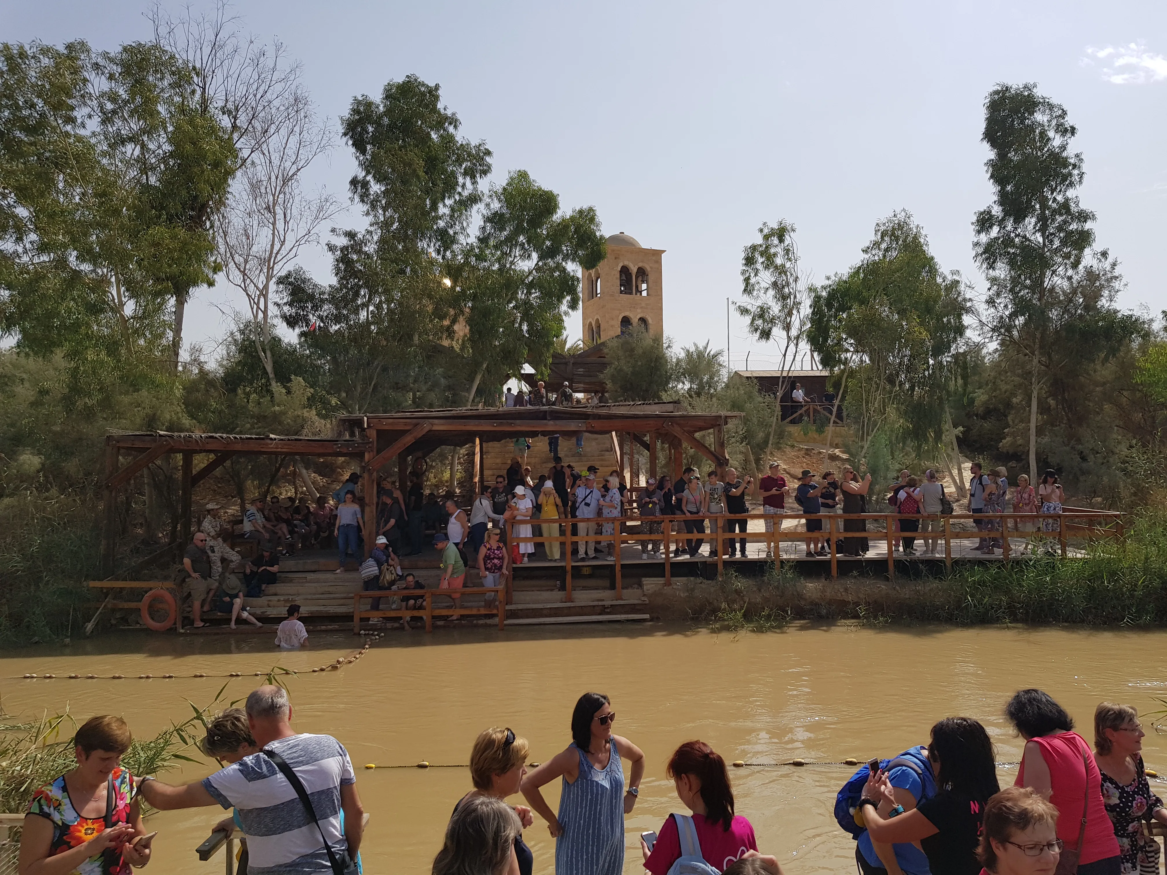 Pilgrims in white robes entering the Jordan River at the Qasr el-Yahud baptism site, with the Jordanian bank visible across the water