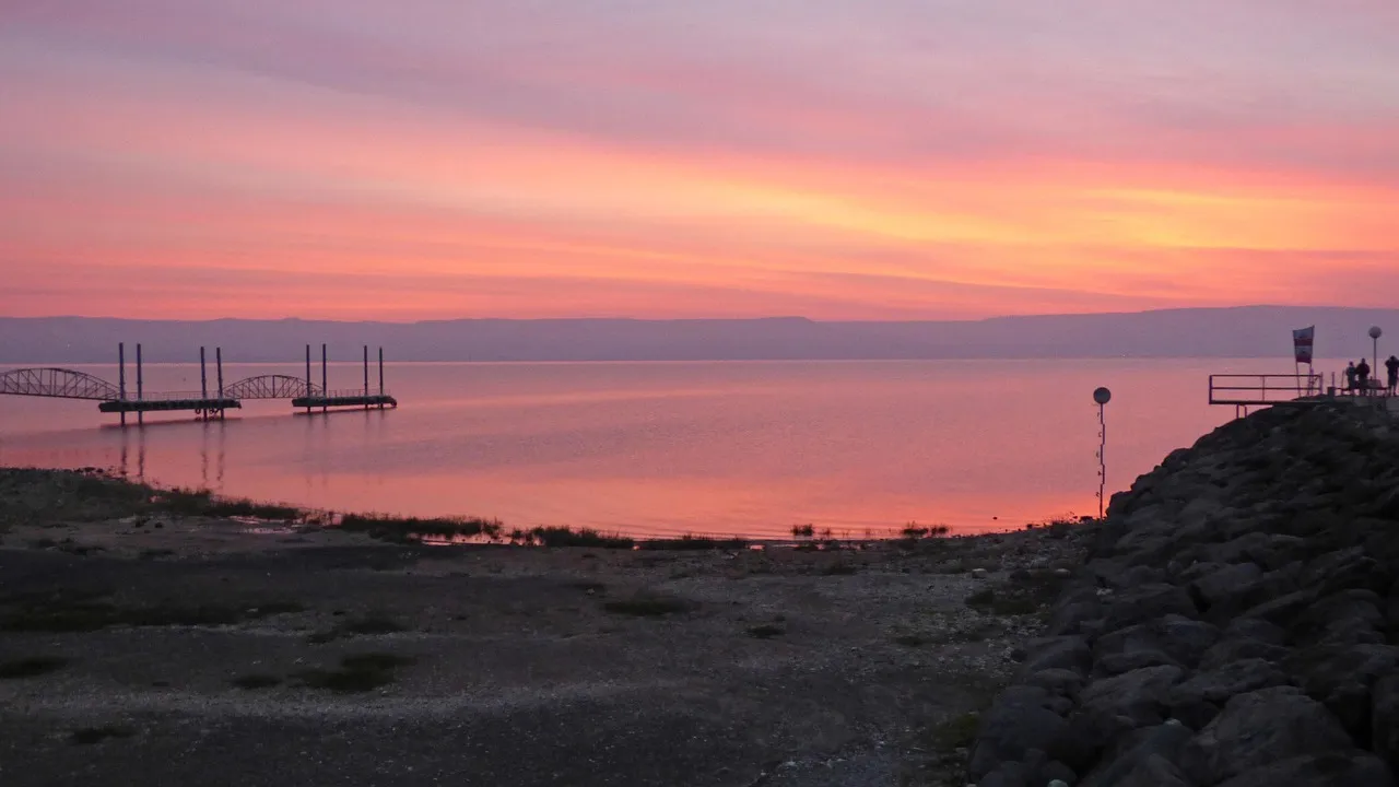 Sea of Galilee at dawn, calm water reflecting golden morning light with the Golan Heights in the distance